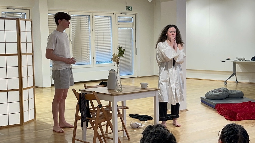 Two actors stand facing the audience in mid-performance of 'Faith Hope Charity'. A table and chairs are between them with a mattress behind them on the right. A few audience members are seen from the back, watching the performance.